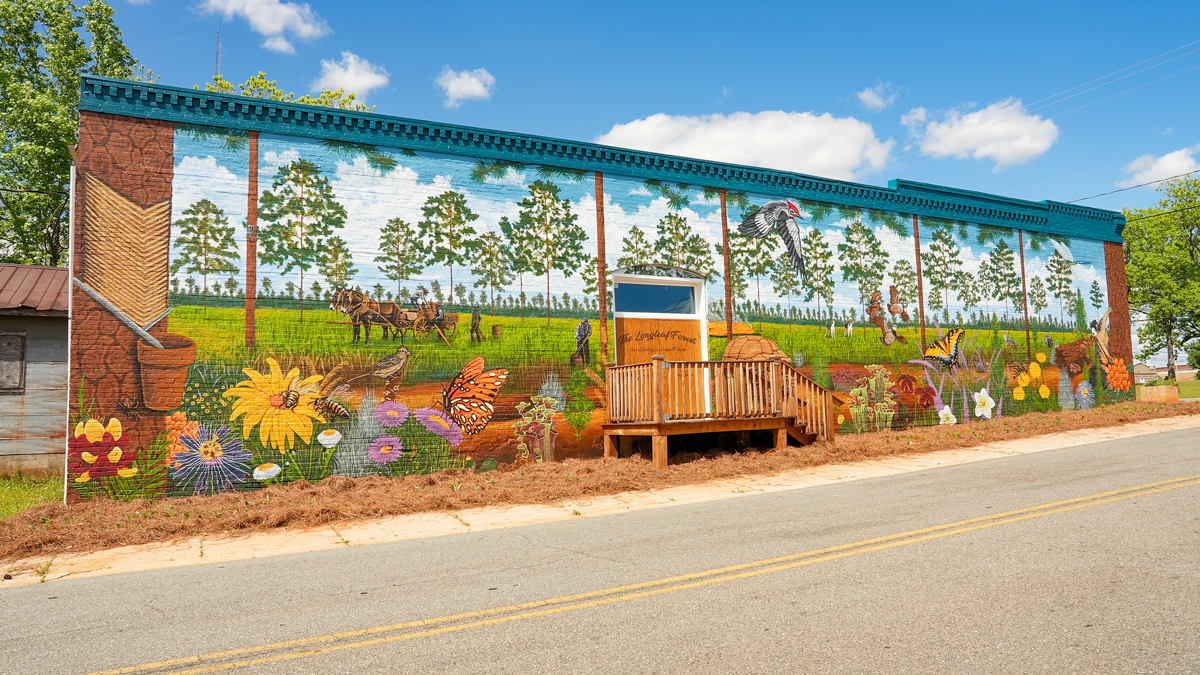 The image depicts a vibrant mural on the side of a building, showcasing a serene natural scene with trees, flowers, and animals, set against a blue sky with white clouds.