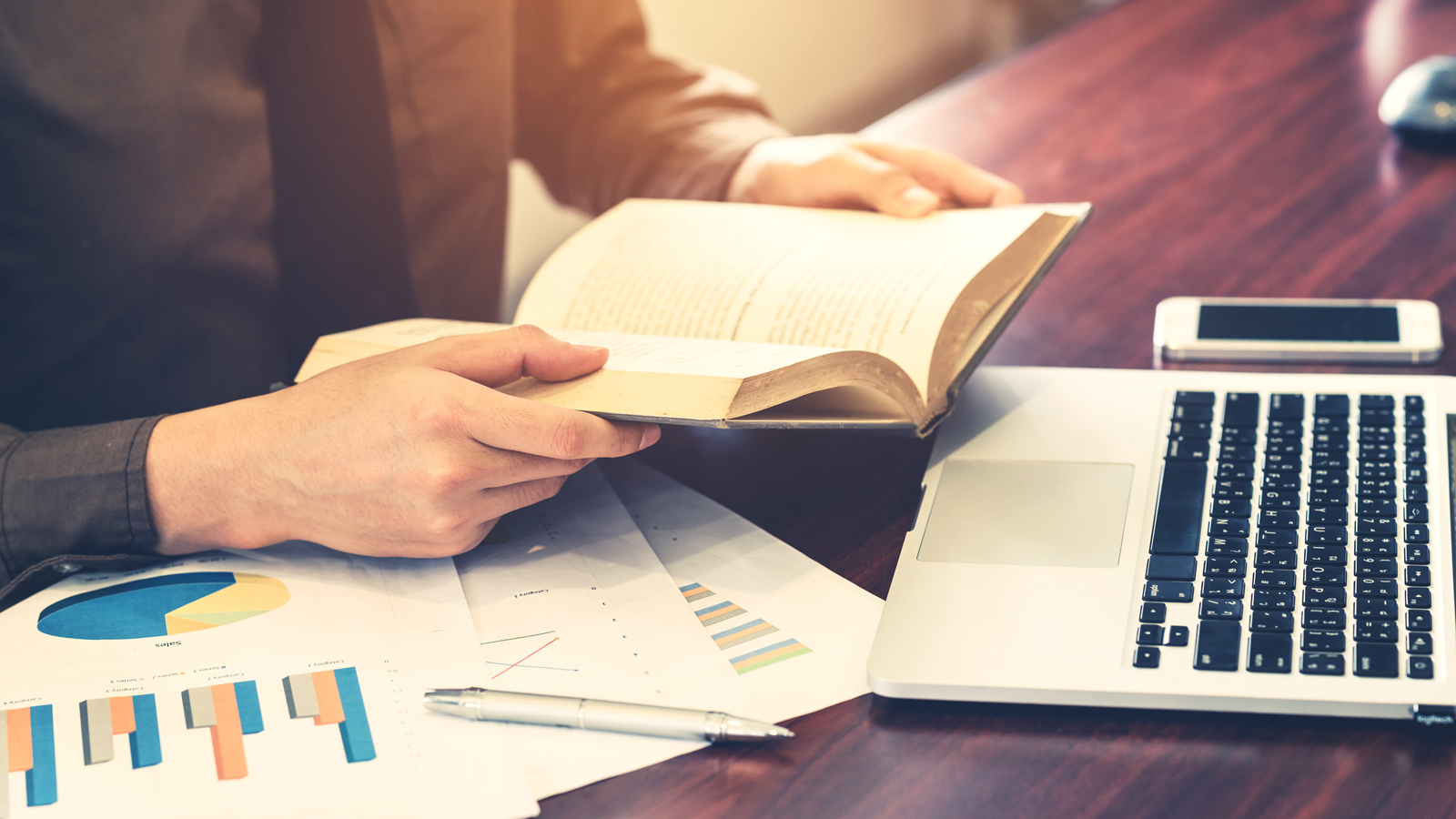 This image shows a person reading a book on a desk with a laptop, cell phone, and papers with charts on them. The person is wearing a tie and brown shirt.