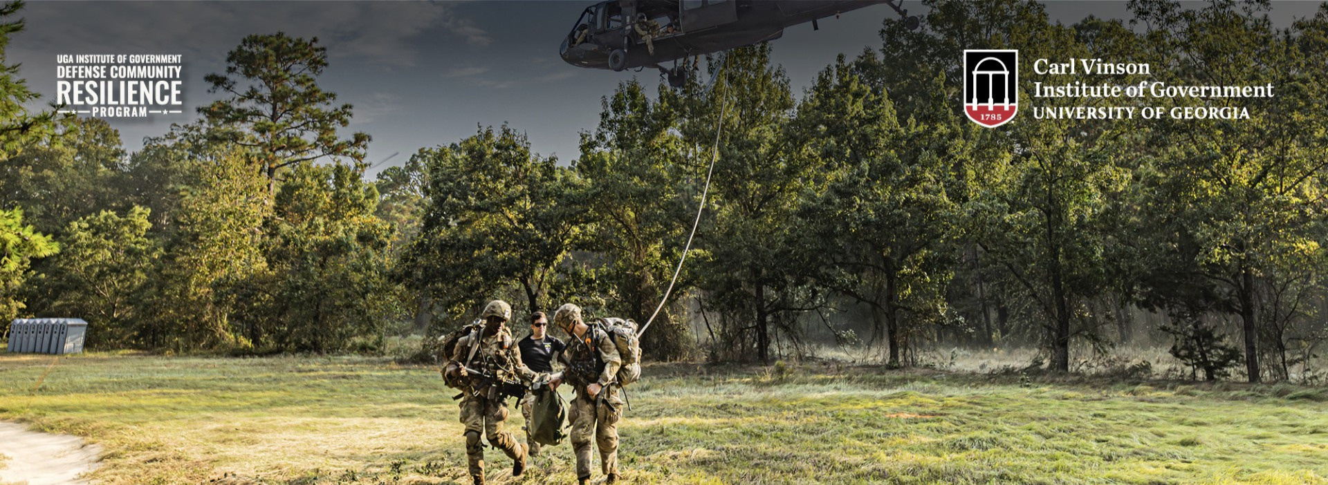 Soldiers carry a person across a grassy field during a helicopter rescue training exercise as a military helicopter hovers overhead.